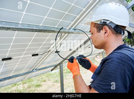 Un travailleur masculin portant un casque de sécurité et des gants de travail tout en câblant les panneaux solaires ensemble.Homme installant un système de panneaux solaires photovoltaïques à l'extérieur.Concept de sources d'énergie renouvelables. Banque D'Images