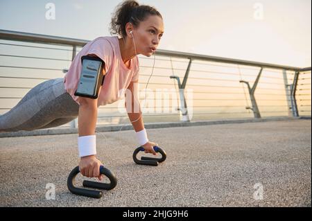 Athlète féminin déterminé, African American sportswoman avec écouteurs et support de smartphone s'entraîner, faire des push-up, tout en exécutant un Banque D'Images