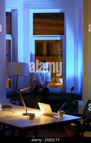 Le Manager ou le propriétaire d'entreprise pensif s'assoit sur le rebord de la fenêtre dans le bureau de nuit en pensant à la solution du problème Banque D'Images