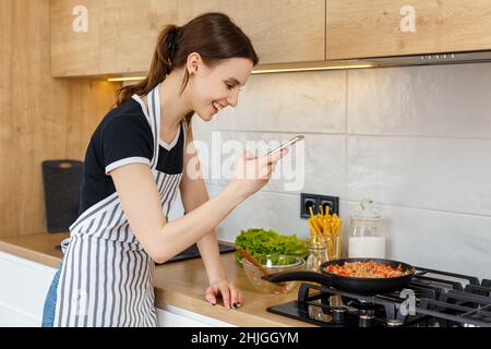 Une jeune femme blogueuse en tablier prend des photos avec le téléphone tout en cuisinant de la nourriture dans la cuisine.Préparation du repas avec une poêle à frire sur une cuisinière à gaz.Concept de style de vie domestique, de loisirs au foyer et de blogging culinaire. Banque D'Images