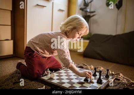 l'enfant joue une seule partie d'échecs à la maison sur le sol.Échiquier avec la fin de la partie d'échecs.La main de la fille se déplaçant une pièce d'échecs est partiellement visible Banque D'Images