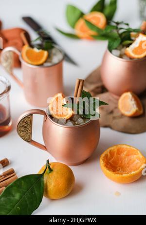 Cocktails glacés à la mandarine d'hiver et à l'alcool bâton de cannelle avec menthe fraîche dans des mugs en cuivre vintage sur fond de table blanc, gros plan Banque D'Images