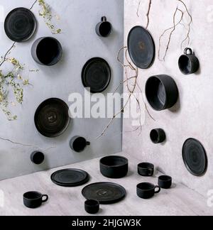 Ensemble de assiettes, bols et tasses en céramique noir artisanal vides en trois dimensions sur table et murs gris, avec des branches sèches comme décoration. Banque D'Images