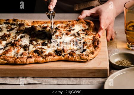 Trancher à la main une pizza aux champignons, oignons et saucisses sur une planche à découper. Banque D'Images