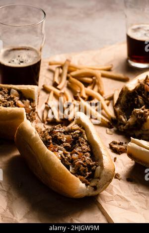 Cheesesteaks et frites sur papier parchemin avec verres de bière foncée. Banque D'Images