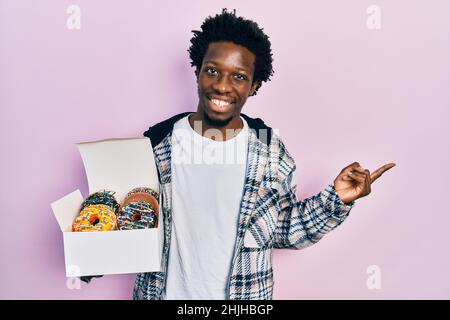 Jeune afro-américain tenant une boîte de beignets colorés et savoureuse, souriante, pointant avec la main et le doigt sur le côté Banque D'Images