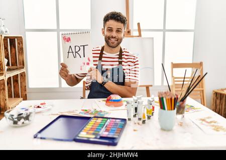 Jeune artiste arabe homme souriant heureux tenant le carnet avec le mot d'art au studio d'art. Banque D'Images