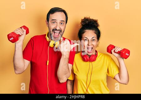 Couple d'âge moyen de femme et d'homme hispaniques portant des vêtements de sport en utilisant des haltères pointant le pouce vers le côté souriant heureux avec la bouche ouverte Banque D'Images