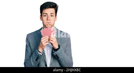 Jeune homme d'affaires hispanique couvrant le visage avec des cartes puffant les joues avec le visage drôle. Bouche gonflée avec de l'air, attrapant l'air. Banque D'Images
