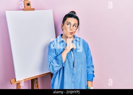 Jeune femme hispanique debout par le chevalet de peintre se concentrant sur le doute avec le doigt sur le menton et regardant vers le haut se demander Banque D'Images