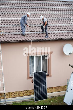 Hommes installateurs de soulever le module solaire photovoltaïque sur le toit de la maison.Électriciens dans les casques d'installation du système de panneaux solaires à l'extérieur.Concept d'énergie alternative et renouvelable. Banque D'Images