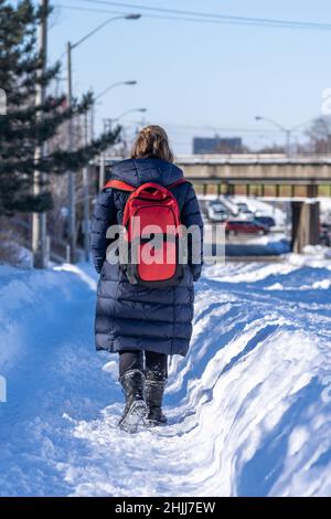 Vue arrière de la jeune femme en vêtement d'extérieur avec sac à dos tenant les mains dans les poches marchant sur un sentier enneigé le jour d'hiver ensoleillé en ville Banque D'Images