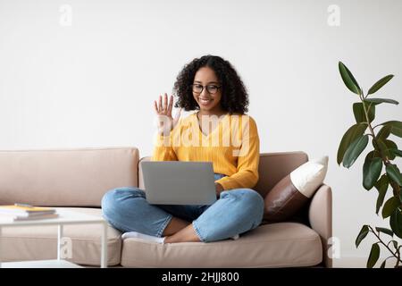 Communication en ligne.Femme noire en lunettes assise sur un canapé à la maison, agitant à la webcam, ayant un appel vidéo à la maison Banque D'Images