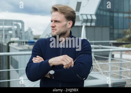 beau blond blanc homme se tient sur le toit et regarde la ville, professionnel, pause au travail, style décontracté homme dans un chandail bleu, directeur Banque D'Images