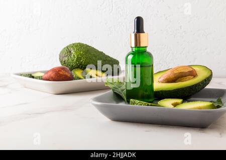 huile cosmétique avec extrait d'avocat dans une bouteille avec compte-gouttes sur une plaque rectangulaire en céramique avec fruits d'avocat contre un mur blanc Banque D'Images