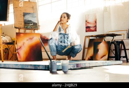 Une jeune artiste s'accroupir sur le sol tout en travaillant dans son studio d'art.Femme peintre regardant la caméra et tenant un pinceau.Jeune wom créatif Banque D'Images