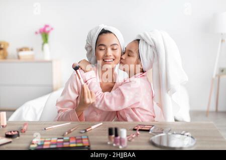 Une adolescente coréenne satisfaite embrasse une jeune femme en pyjama rose et une serviette sur la tête à table avec des cosmétiques Banque D'Images