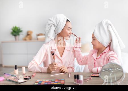 Une jeune fille chinoise gaie et une jeune femme en pyjama rose et des serviettes sur la tête font du maquillage dans la chambre Banque D'Images