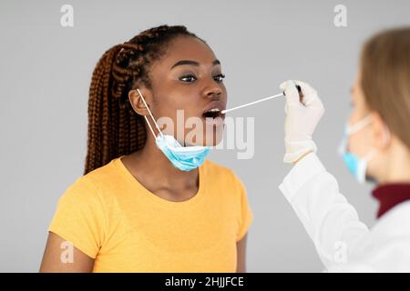 Médecin effectuant un test PCR par coronavirus oral pour une patiente noire sur fond gris.Protection contre les maladies infectieuses Banque D'Images