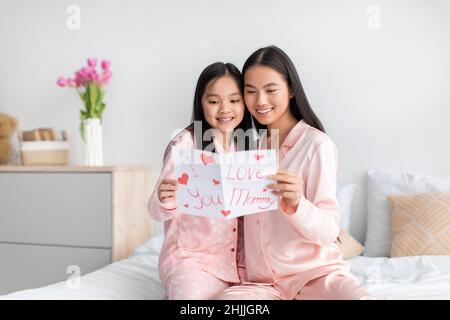 Bonne fille asiatique et jeune femme lisant une carte postale assise sur un lit dans un intérieur minimaliste Banque D'Images