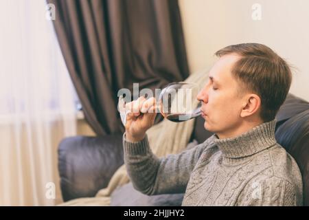 Décontracté, serein, riche, solitaire, d'âge moyen, un homme seul boit du vin rouge, se détendant sur un canapé à la maison, se reposant sur un canapé tenant une dégustation de verre Banque D'Images