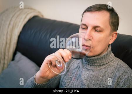 Décontracté, serein, riche, solitaire, d'âge moyen, un homme seul boit du vin rouge, se détendant sur un canapé à la maison, se reposant sur un canapé tenant une dégustation de verre Banque D'Images