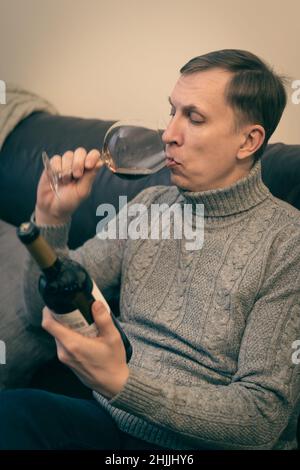 Décontracté, serein, riche, solitaire, d'âge moyen, un homme seul boit du vin rouge, se détendant sur un canapé à la maison, se reposant sur un canapé tenant une dégustation de verre Banque D'Images