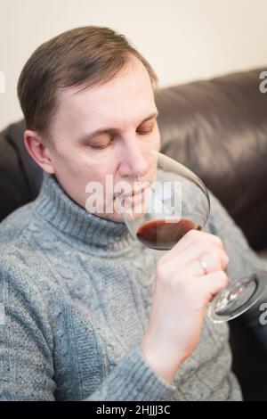 Décontracté, serein, riche, solitaire, d'âge moyen, un homme seul boit du vin rouge, se détendant sur un canapé à la maison, se reposant sur un canapé tenant une dégustation de verre Banque D'Images