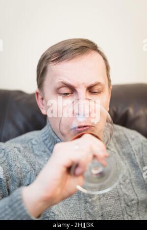 Décontracté, serein, riche, solitaire, d'âge moyen, un homme seul boit du vin rouge, se détendant sur un canapé à la maison, se reposant sur un canapé tenant une dégustation de verre Banque D'Images