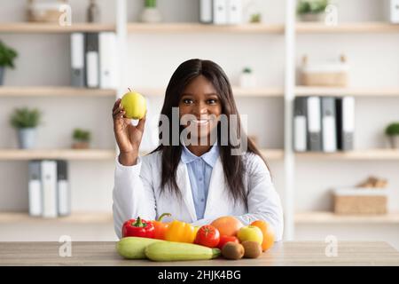 Portrait d'une diététiste noire tenant la pomme, recommandant des fruits et légumes frais, assis dans un bureau médical Banque D'Images