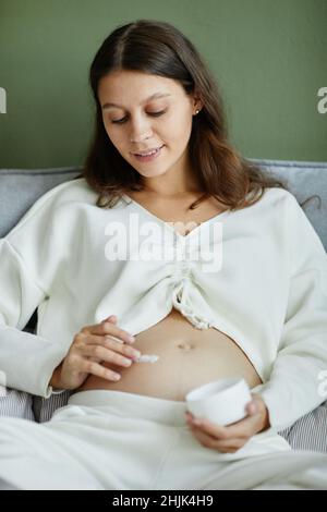 Jeune femme enceinte s'occupant de sa peau pendant la grossesse, elle applique de la crème cosmétique sur son ventre allongé dans le lit Banque D'Images