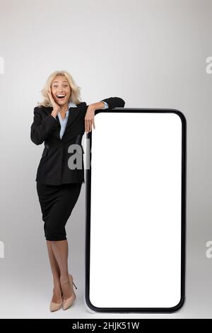 Application professionnelle.Femme entrepreneur enthousiaste en tenue formelle penchée au grand smartphone blanc avec écran blanc, maquette Banque D'Images