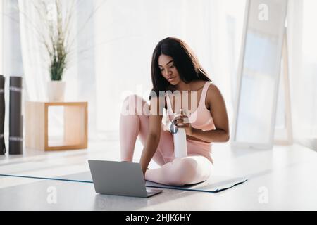 Jolie jeune femme noire en vêtements de sport assise sur un tapis de yoga, en utilisant un ordinateur portable, tenant une bouteille d'eau à l'intérieur, sur toute la longueur Banque D'Images