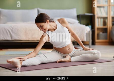 Bonne jeune femme enceinte assise sur un tapis d'exercice et s'étirant pendant l'entraînement sportif dans la chambre à la maison Banque D'Images