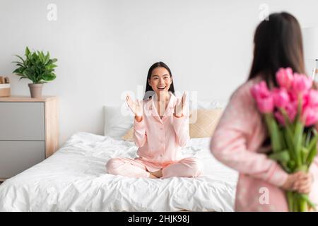 Jeune fille asiatique avec bouquet de tulipes, dos et bonne jeune femme assise sur le lit dans la chambre à coucher à l'intérieur Banque D'Images