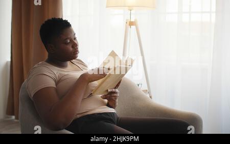Concept d'auto-éducation.Jeune femme afro-américaine concentrée qui lit un livre intéressant, reposant sur un fauteuil Banque D'Images