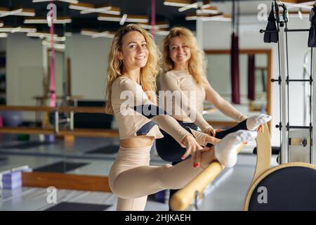 Deux femmes de fitness souriant à la caméra tout en s'étirant dans le studio pilates moderne dans la journée.Sports, mode de vie sain, concept d'amitié Banque D'Images