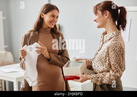 Bonne jeune femme enceinte regardant des vêtements de bébé donner par son collègue, ils parlent debout au bureau Banque D'Images