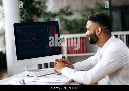 Smart Indian mâle crypto Trader, investisseur, regardant l'écran d'ordinateur, analyser les diagrammes de négociation sur le marché boursier d'une bourse de crypto-monnaie numérique, achète et vend de la crypto-monnaie, analyse dynamique Banque D'Images