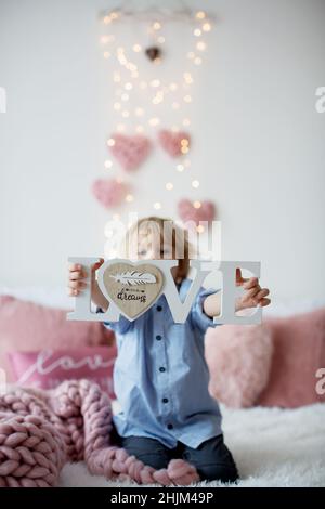 Mignon enfant blond avec chemise, assis sur le lit avec décoration de Saint-Valentin, arrière-plan isolé Banque D'Images