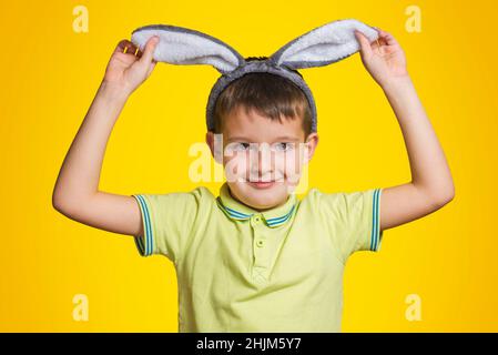 Le petit garçon en t-shirt touche ses oreilles de lapin et sourit tout en se tenant sur fond jaune.Petit enfant mignon portant des oreilles de lapin le jour de Pâques. Banque D'Images