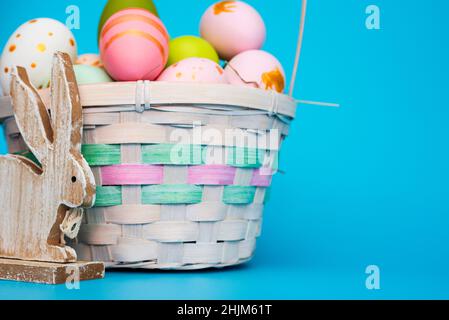Un panier d'œufs de Pâques avec lapin sur fond bleu.Panier festif avec œufs peints en couleur et lapin en bois jouet.Composition de Pâques. Banque D'Images