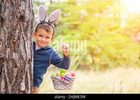 Un petit garçon mignon avec des oreilles de lapin debout derrière un arbre et tenant un panier rempli d'oeufs de pâques colorés après la chasse aux oeufs dans sa main.Joyeuses Pâques Banque D'Images