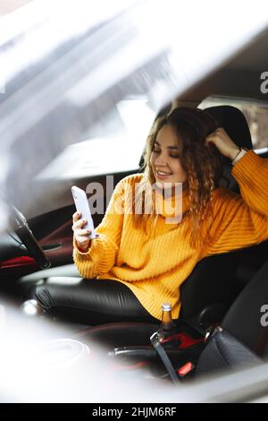 Portrait d'une jeune femme décontractée dans un chandail orange, textant sur son smartphone ou faisant un appel vidéo, tout en conduisant ou en faisant un arrêt de circulation une voiture pendant la pause déjeuner. Banque D'Images