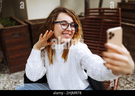 Bonne jeune caucasienne, millénaire ou la femme de génération z avec de longs cheveux de brunette streaming avec le smartphone en main, le tournage de blog de médias sociaux dans le café extérieur moderne.Hipster Influencer en utilisant les réseaux sociaux. Banque D'Images