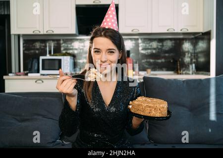 Fille mangeant gâteau assis sur le canapé à la maison, regardant l'appareil photo, la femme heureuse a le dessert, la nourriture de junk et étant en surpoids, la nourriture de vacances Banque D'Images