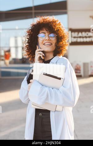 Femme entrepreneur avec ordinateur portable parlant au téléphone tout en marchant le long de la rue ensoleillée.Femme d'affaires aux cheveux bouclés.Employé de bureau.Communication mobile Banque D'Images