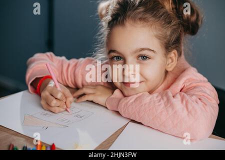 Portrait en gros plan d'une petite fille caucasienne en dessinant dans un bloc-notes avec des stylos colorés regardant l'appareil photo et assis au bureau à la maison.Mode de vie de quarantaine Banque D'Images