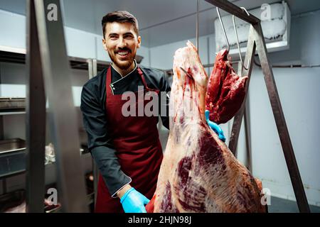 Boucherie mâle debout près de la viande accrochée au crochet dans la boutique de viande Banque D'Images