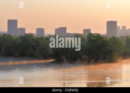 Maisons et arbres sur la rive de la rivière.Grandes maisons, arbres luxuriants sur la rive à l'aube.Brouillard sur l'eau, reflets dans l'eau, couleur rose.Novosi Banque D'Images